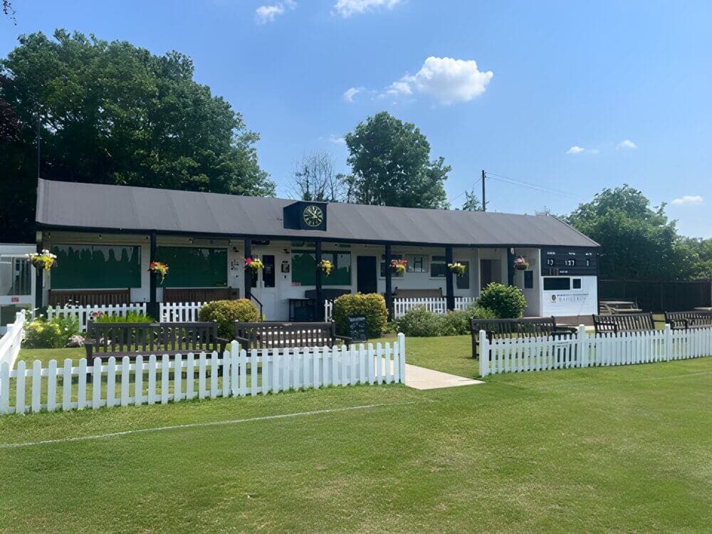 A lawn bowling club with a black roof, white picket fence, green lawn, and trees in the background on a sunny day. - Home Instead