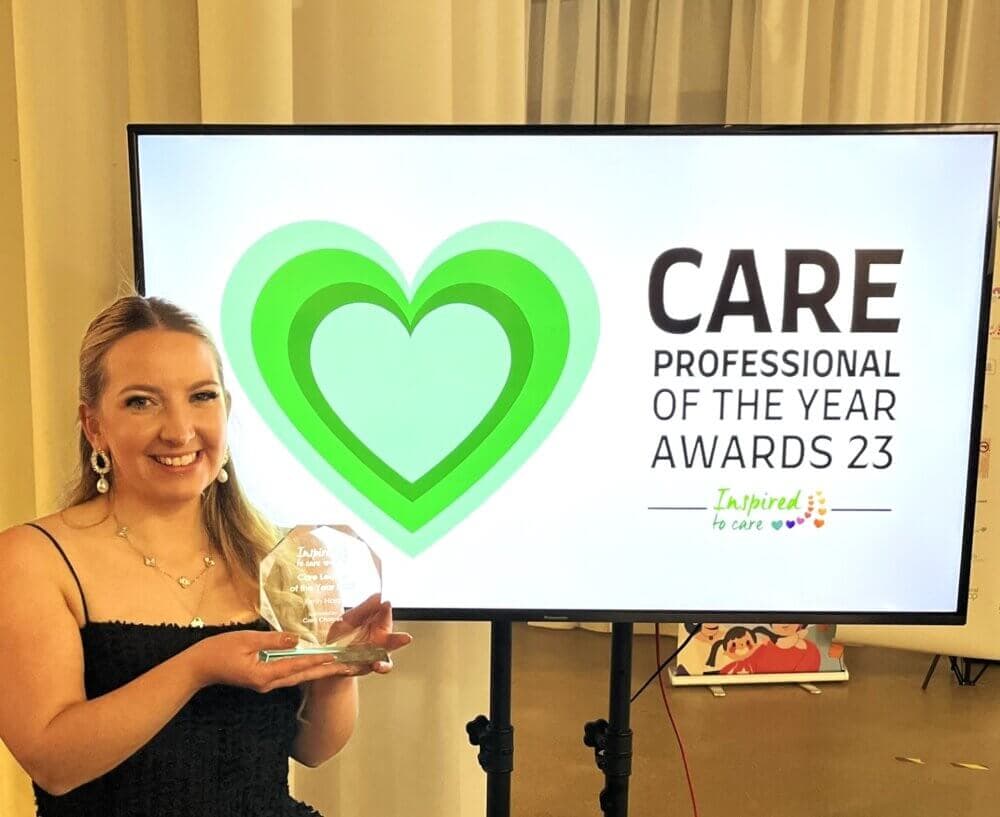 Person smiling and holding an award, standing next to a display reading "Care Professional of the Year Awards 23. - Home Instead