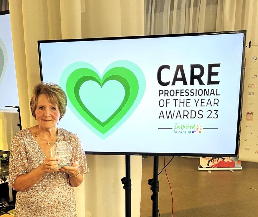 A person holding a trophy stands beside a screen displaying "CARE Professional of the Year Awards 23. - Home Instead