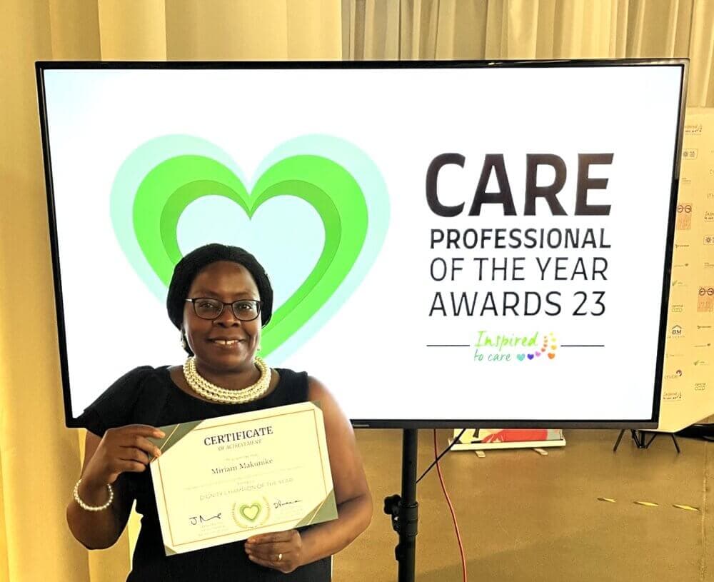 A person holding a certificate stands in front of a screen displaying "Care Professional of the Year Awards 23. - Home Instead
