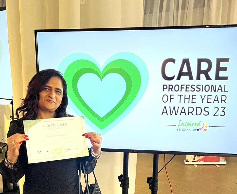 A woman holding a certificate stands in front of a screen displaying "Care Professional of the Year Awards 23. - Home Instead