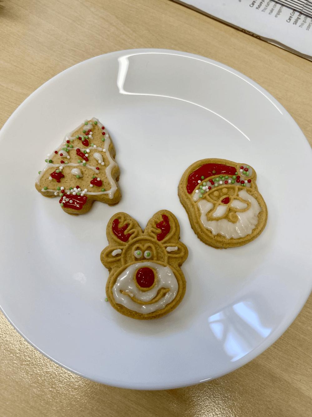 A white plate on a table containing three Christmas biscuits, shaped as a Christmas tree, Rudolph the red nosed reindeer and Santa Claus - Home Instead