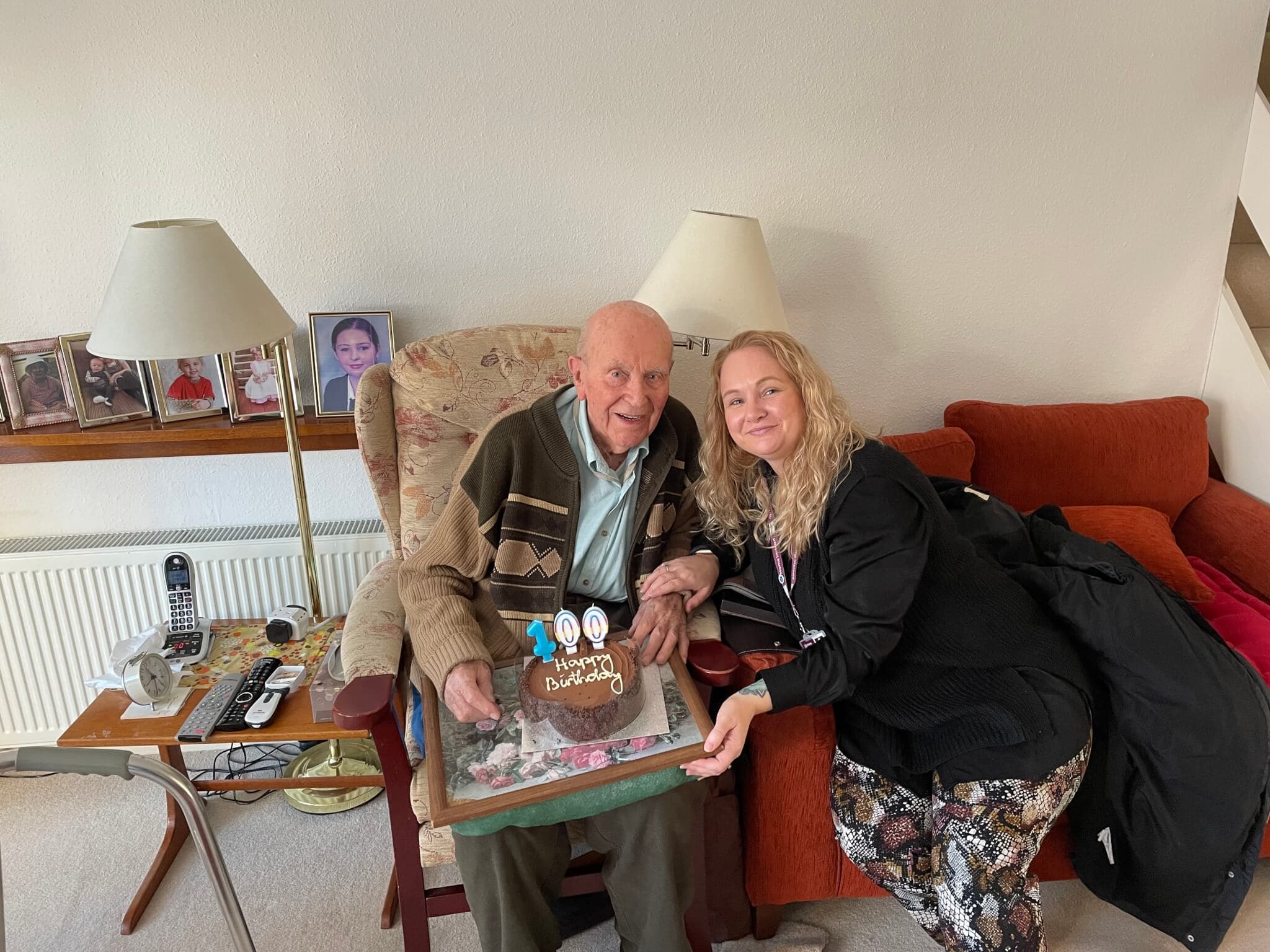 An elderly man celebrates his 100th birthday with a woman, holding a card and cake. - Home Instead