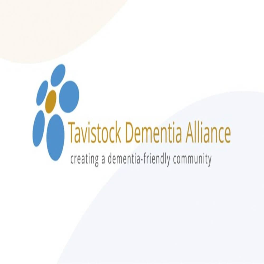 Logo of Tavistock Dementia Alliance with the text "creating a dementia-friendly community" and a blue and orange floral design. - Home Instead