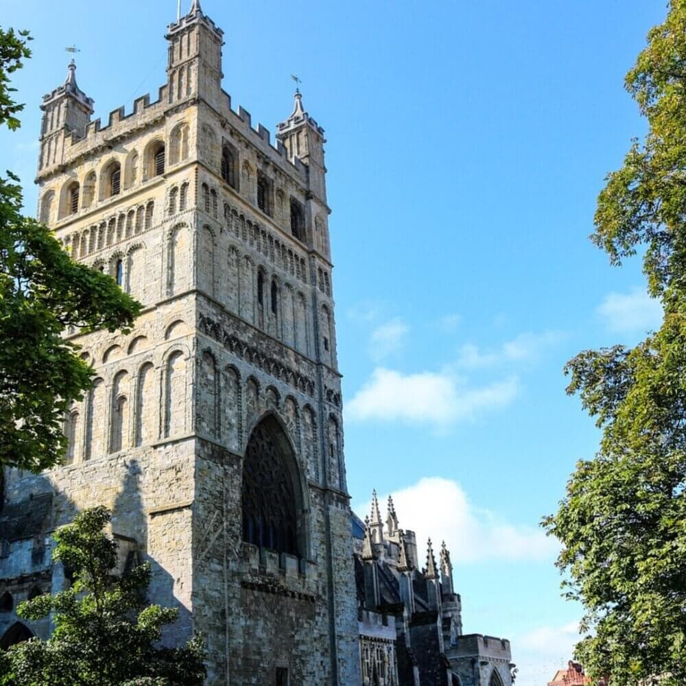 Tall stone cathedral tower with arched windows, surrounded by trees, under a clear blue sky. - Home Instead