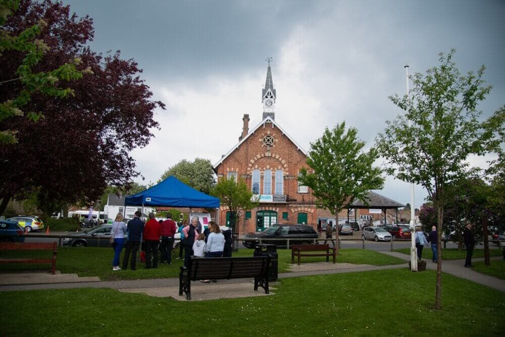 People gather under a blue tent near a red brick building with a clock tower on a cloudy day. - Home Instead