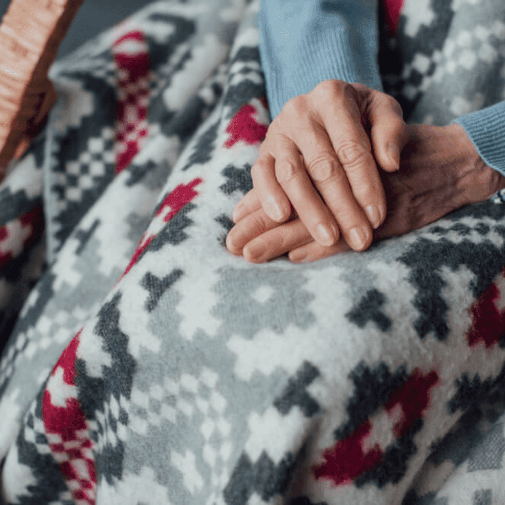 Close-up of an elderly person's hands resting on a patterned, knitted blanket. - Home Instead