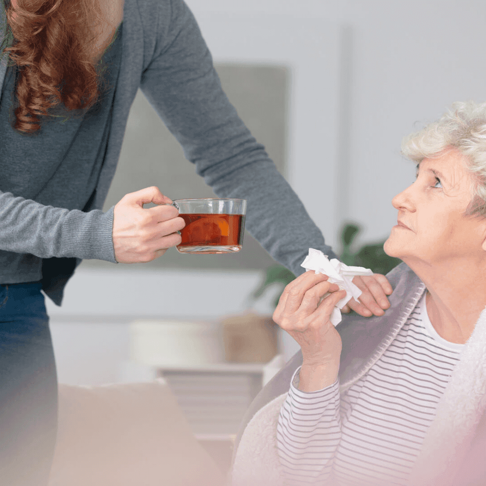 A person offers tea to an older woman holding tissues, while she appears to be unwell. - Home Instead