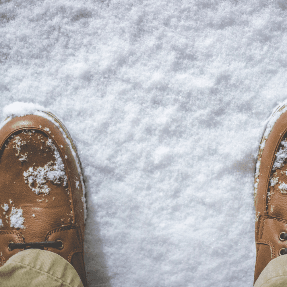 Brown shoes dusted with snow, standing on snow-covered ground, with beige pants partially visible. - Home Instead