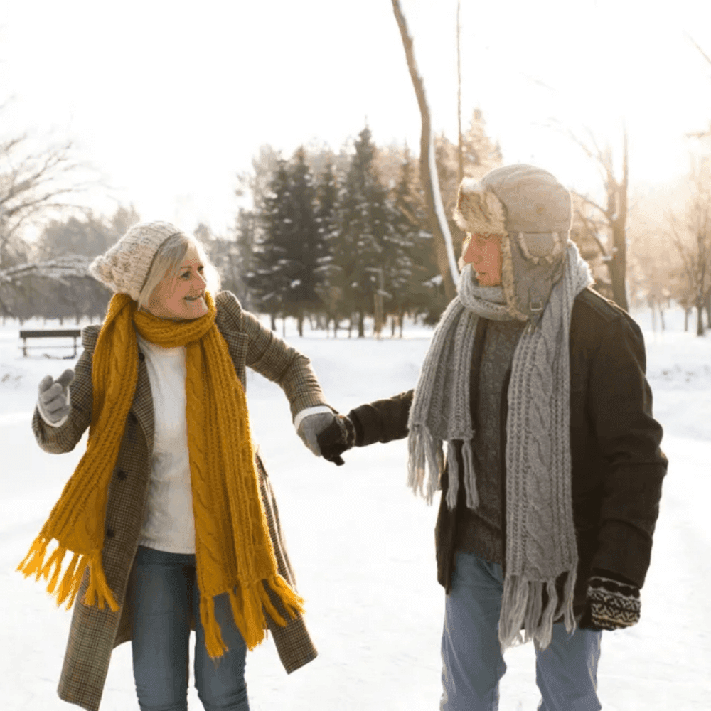 An elderly couple bundled up in winter clothing walk hand in hand through a snowy park, smiling at each other. - Home Instead