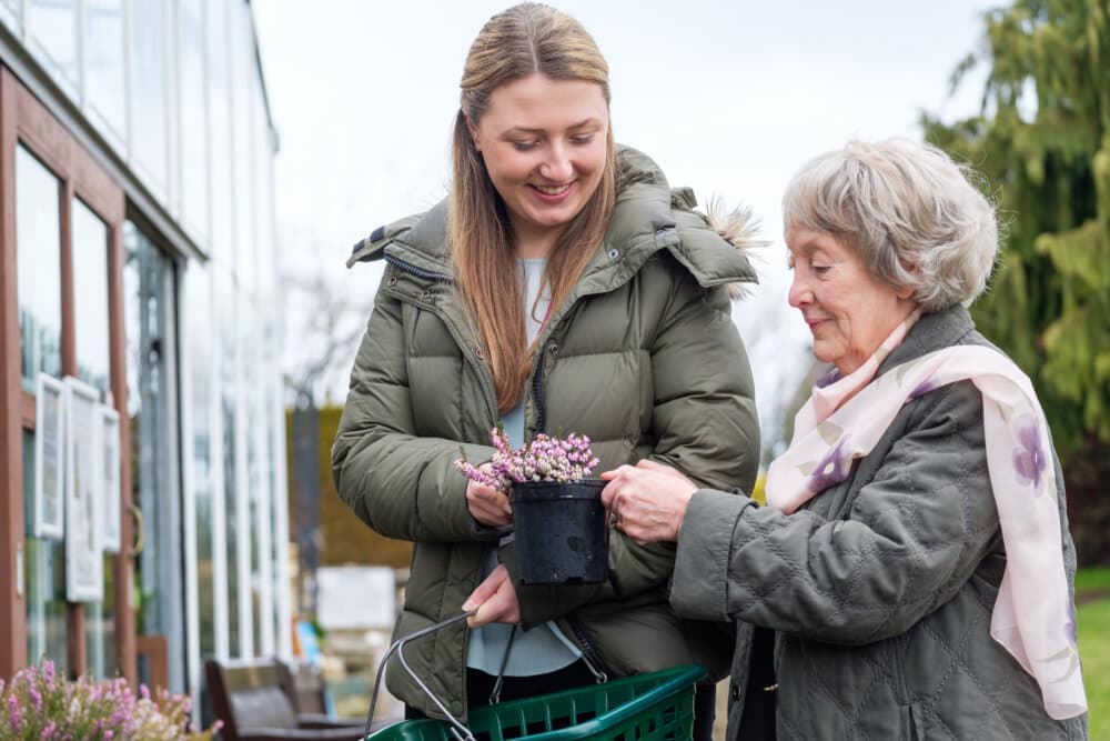 Two women, one elderly, smile while looking at a potted plant outside a greenhouse on a chilly day. - Home Instead