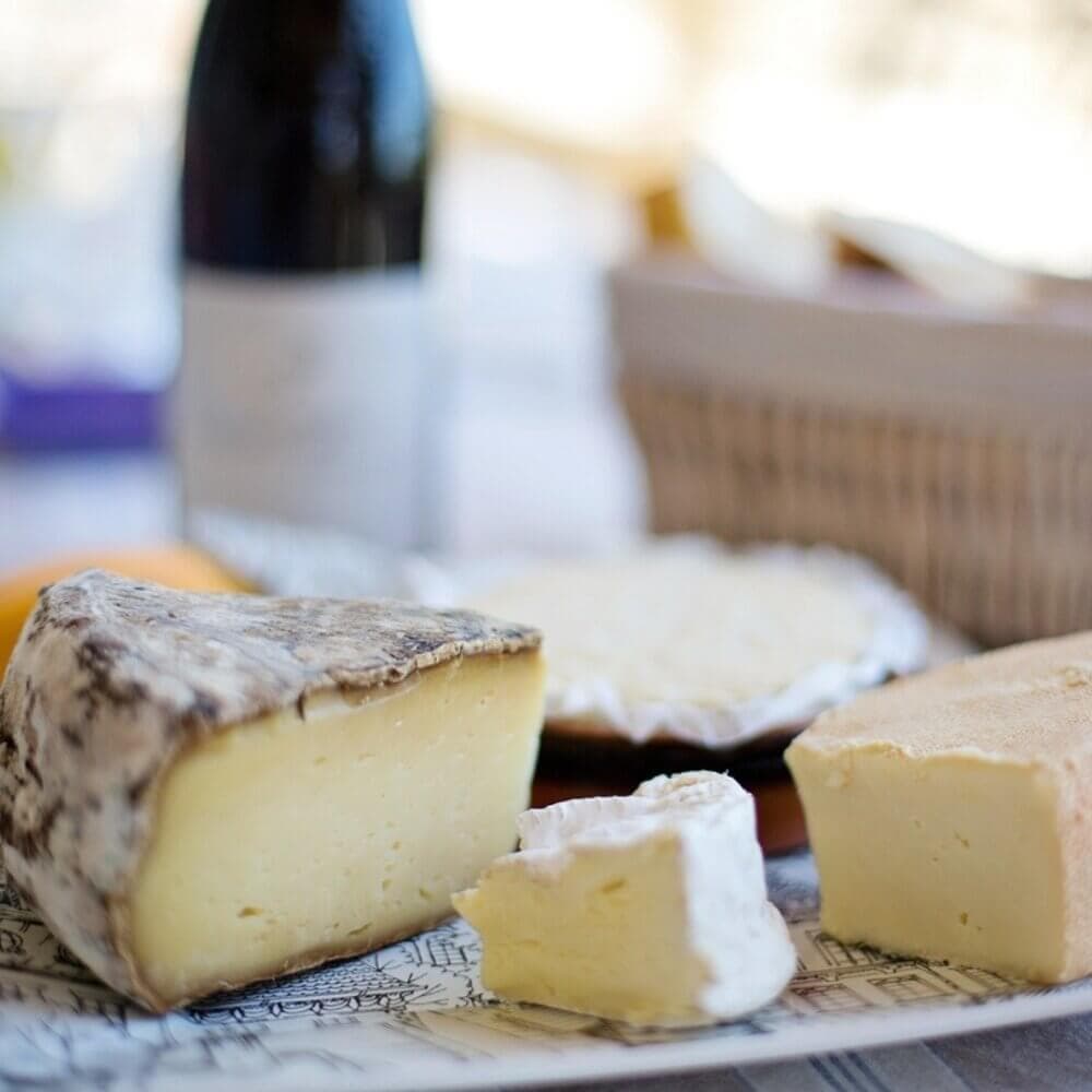 A variety of cheeses and a bottle of wine on a table, with a wicker basket in the background. - Home Instead