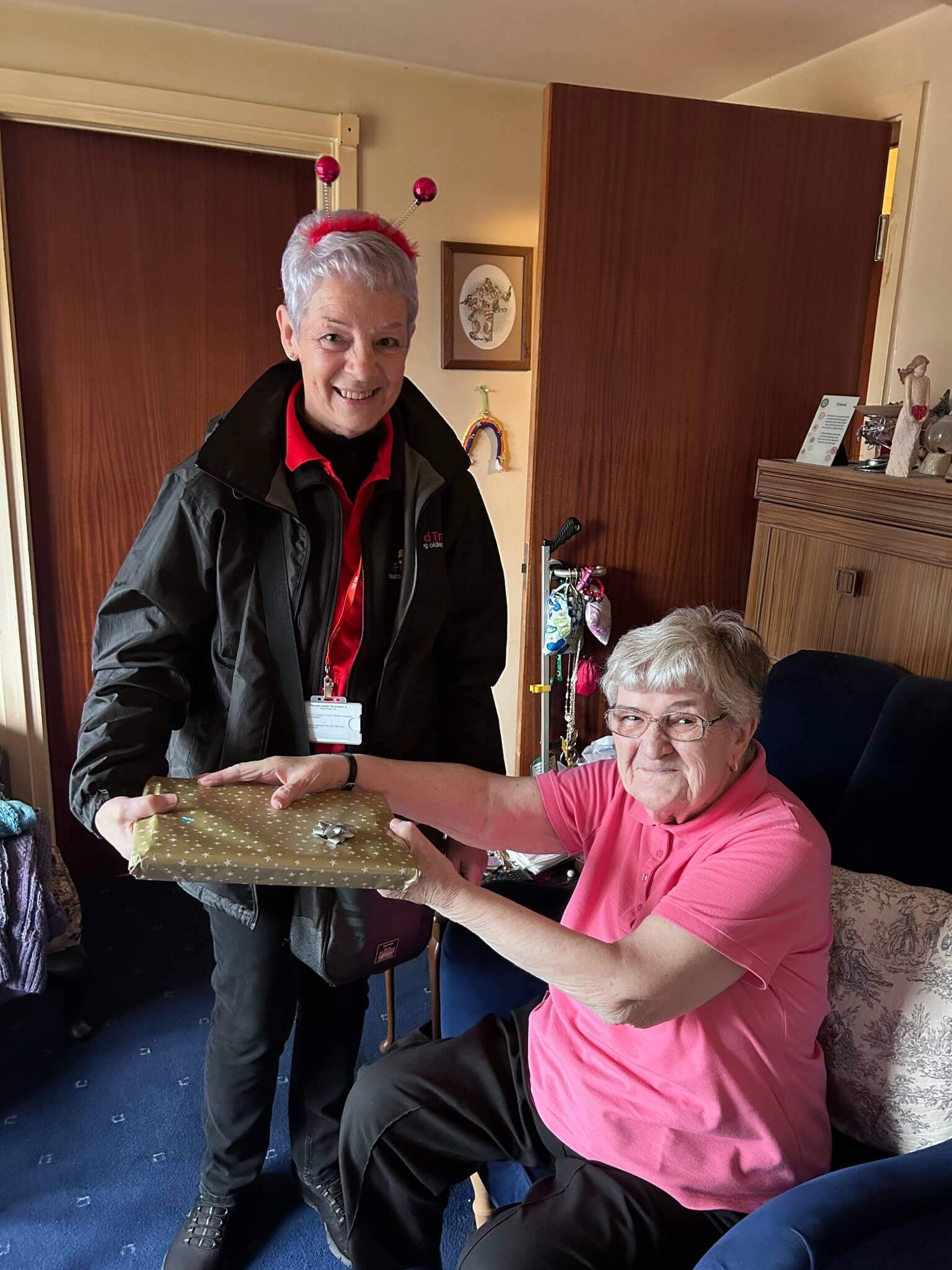 An older woman receives a wrapped gift from a smiling woman in a room with wooden doors and various items displayed. - Home Instead