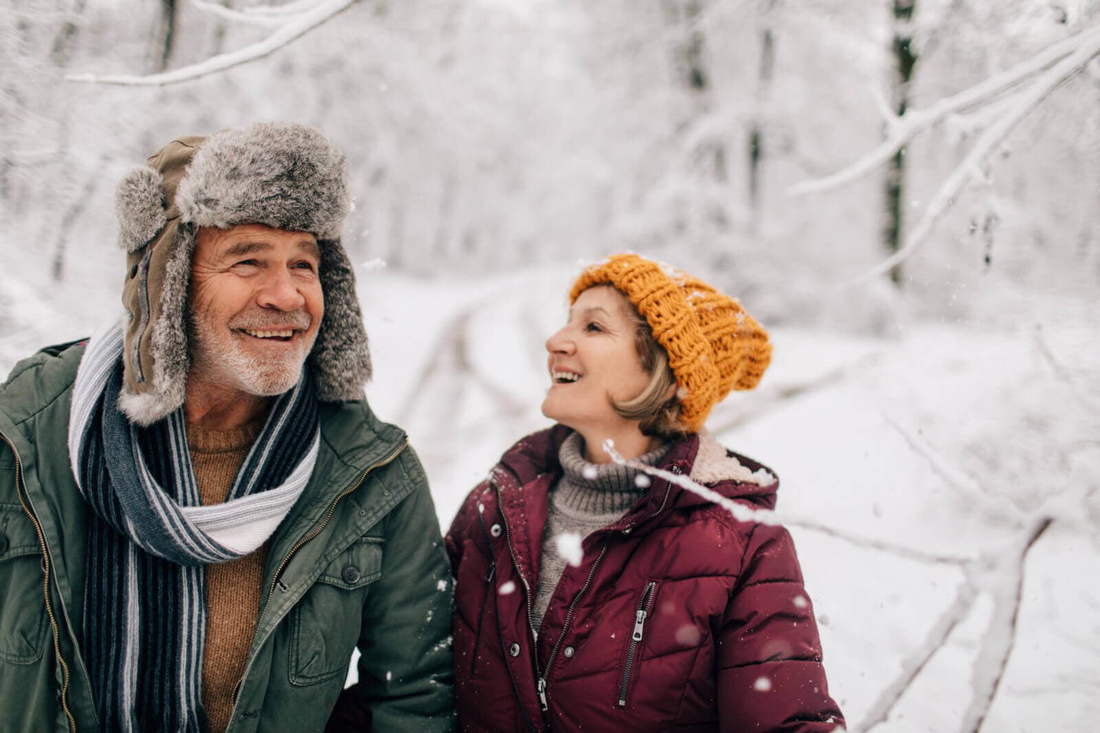An elderly couple in winter attire smiles while walking through a snowy forest. - Home Instead
