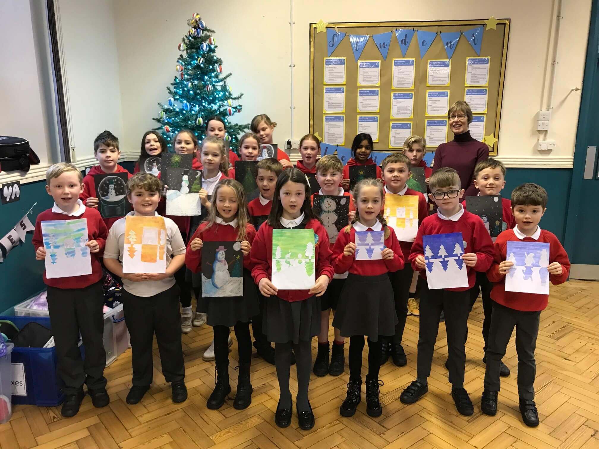 A group of school children proudly holding their artwork, standing with a teacher in front of a decorated Christmas tree. - Home Instead