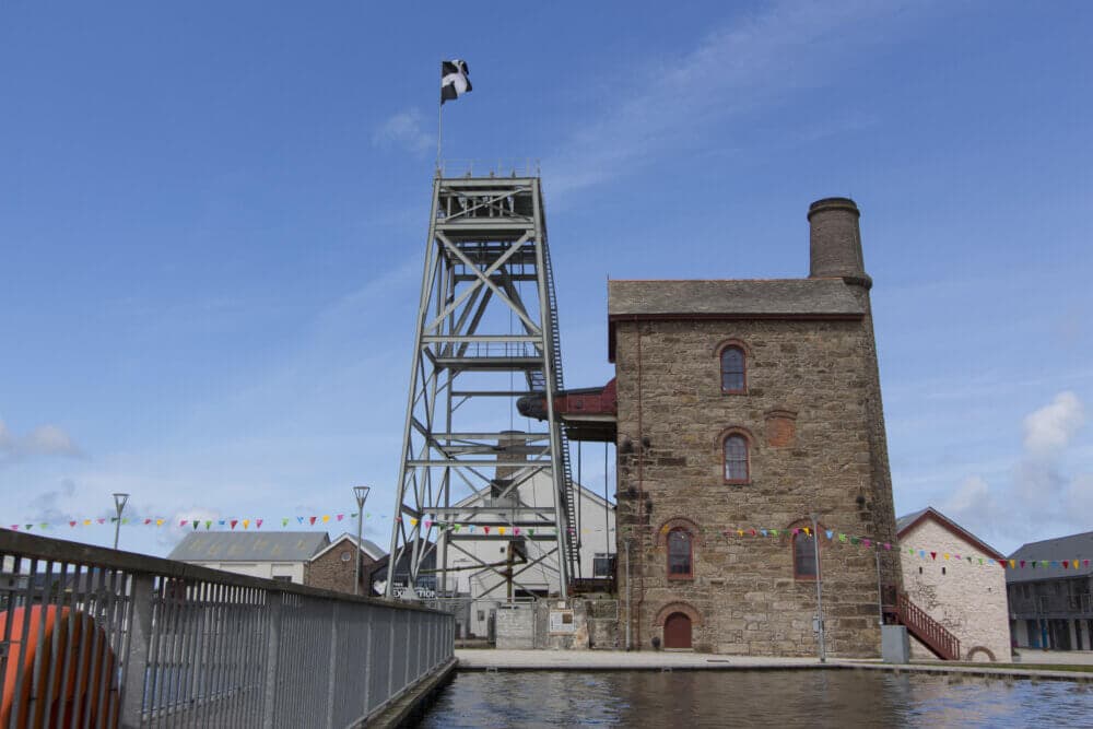 A historical mining building with a tall metal structure and Cornish flag under a clear sky. Colorful bunting is displayed. - Home Instead