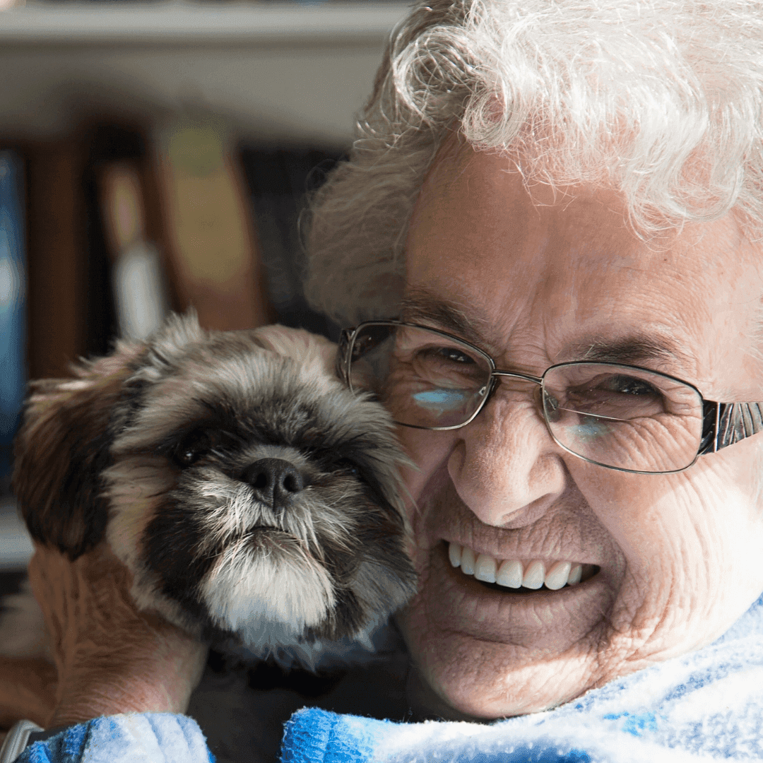 An elderly person with glasses smiles while holding a small dog close to their face. Both appear happy and content. - Home Instead