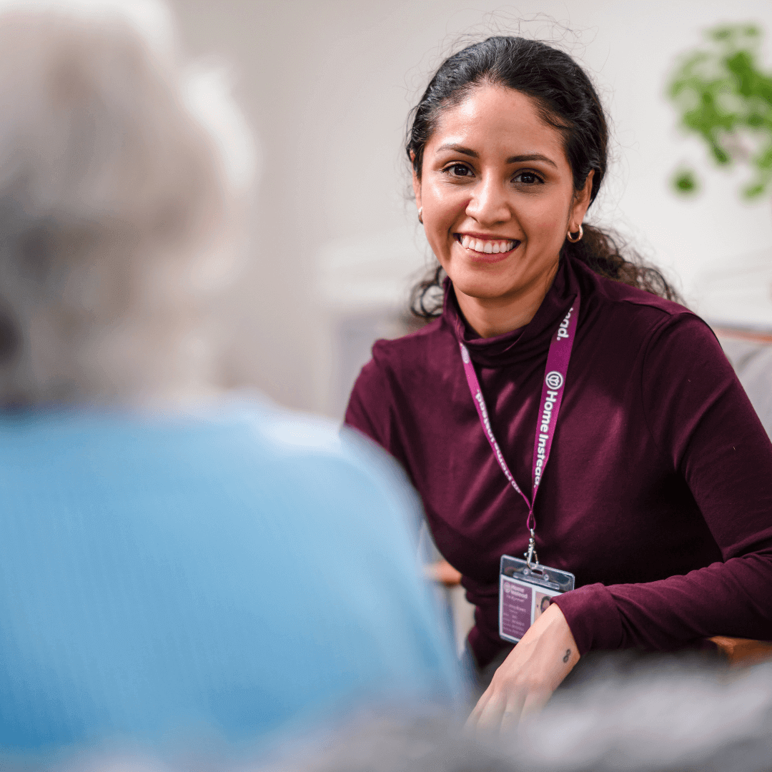 A caregiver in a maroon shirt and name badge smiling at an elderly person, who is blurred in the foreground. - Home Instead