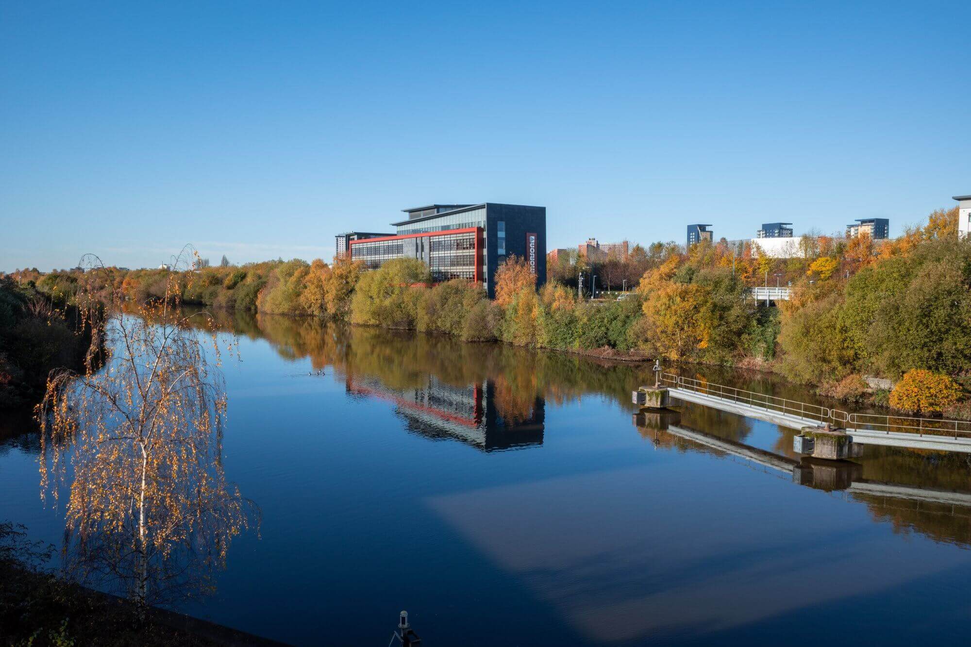 A calm river reflecting a modern building and autumn trees on a clear day with blue sky. - Home Instead