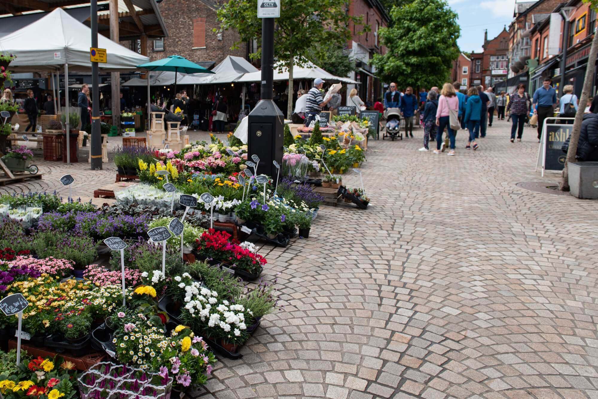 A market street with flower stalls and various goods. People are browsing and walking around on a cobblestone pathway. - Home Instead