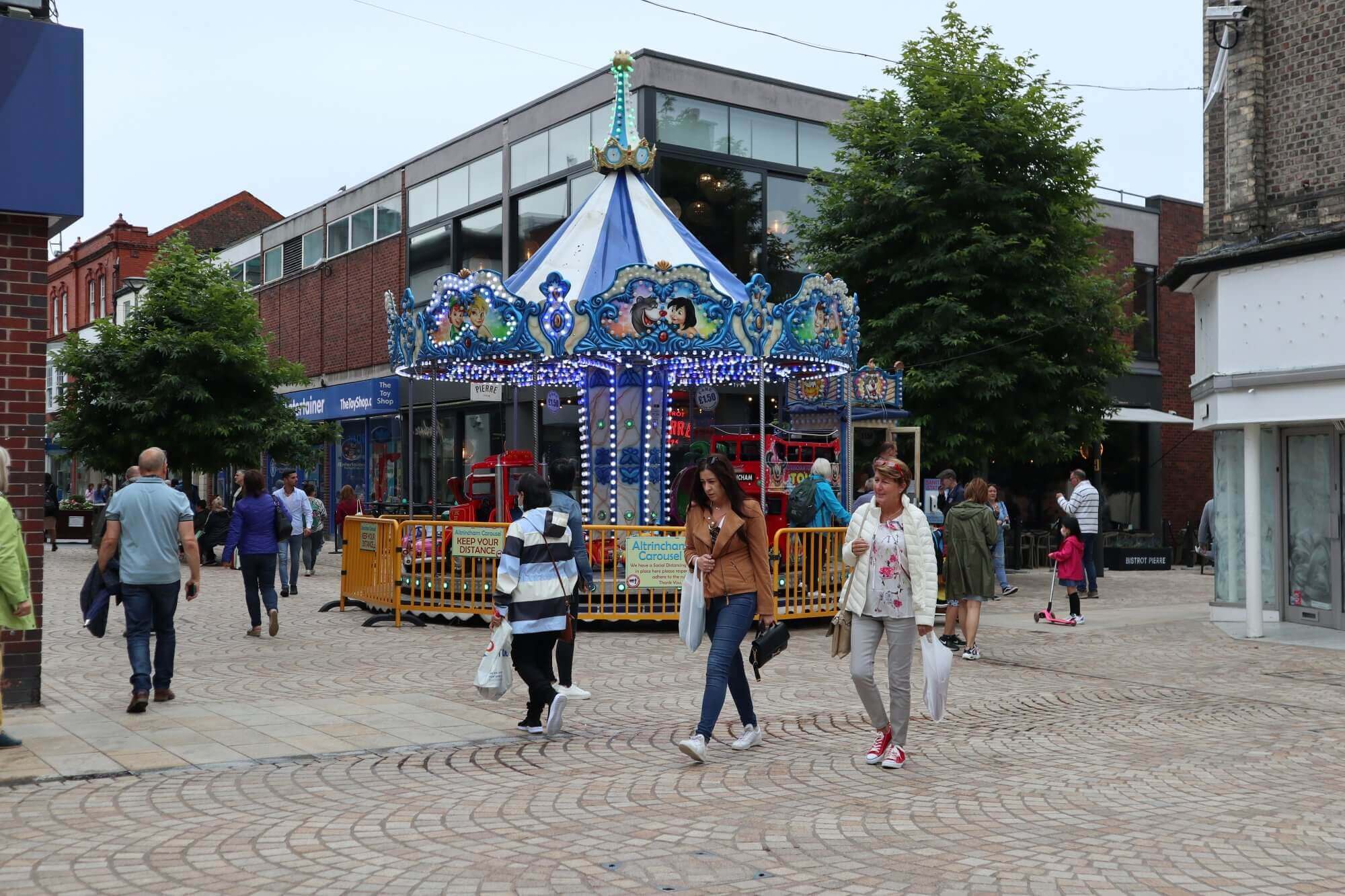 People walking near a carousel on a cobblestone street with shops and trees in the background. - Home Instead
