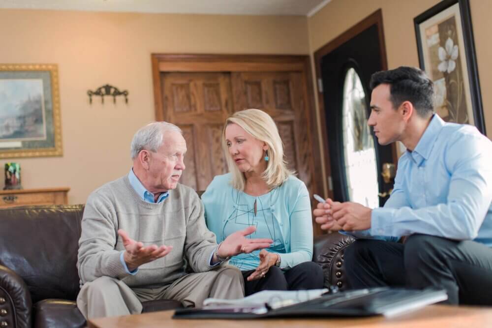 An elderly man and a woman talking to a younger man holding a pen, sitting on a couch in a cozy living room. - Home Instead