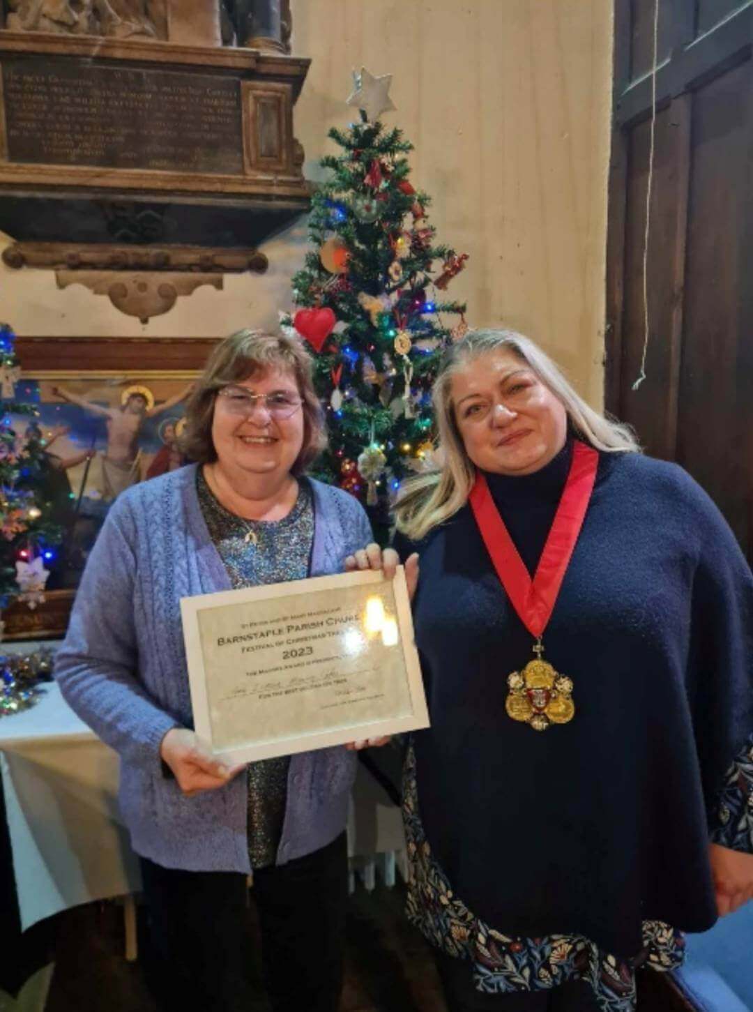 Two women standing in front of a Christmas tree, one holding an award certificate and the other wearing a medal. - Home Instead