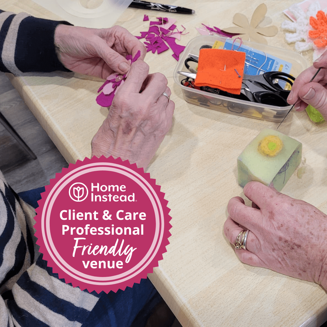 Two people engage in art and craft activities at a table with a "Home Instead: Client & Care Professional Friendly Venue" badge. - Home Instead
