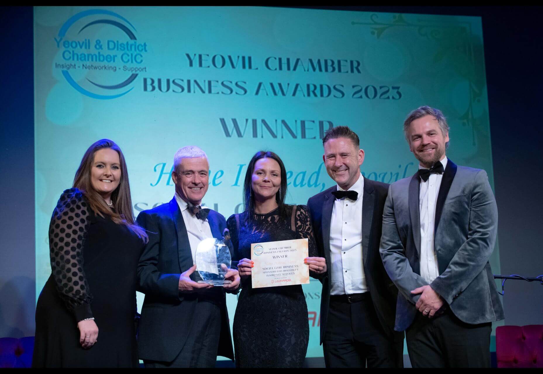 Five people in formal attire on stage at Yeovil Chamber Business Awards 2023; one holding a trophy and another a certificate. - Home Instead