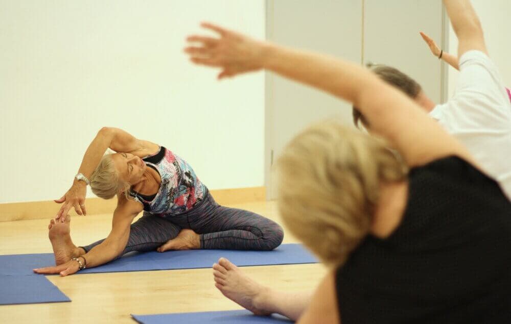 People practicing a seated side stretch in a yoga class, with one instructor guiding them. - Home Instead