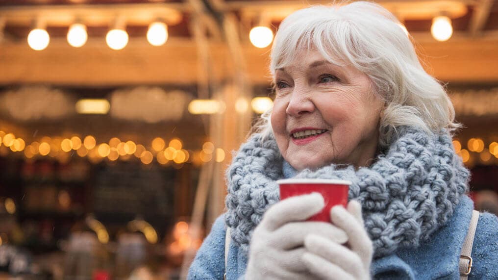 Elderly woman in winter attire smiles while holding a red cup, festive lights glow in the background. - Home Instead