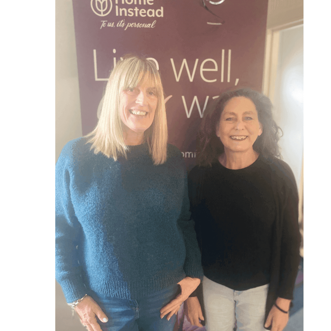 Two smiling women standing in front of a "Home Instead" banner with the text "Live well, your way. - Home Instead