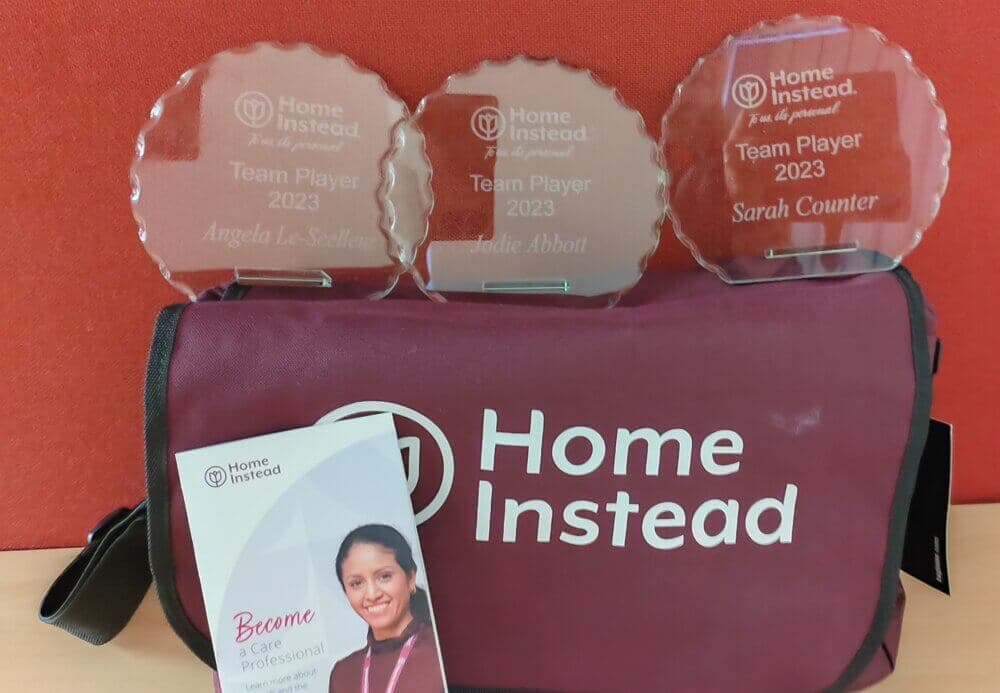 Three "Team Player 2023" awards on a Home Instead bag, with a brochure featuring a smiling woman in the foreground. - Home Instead