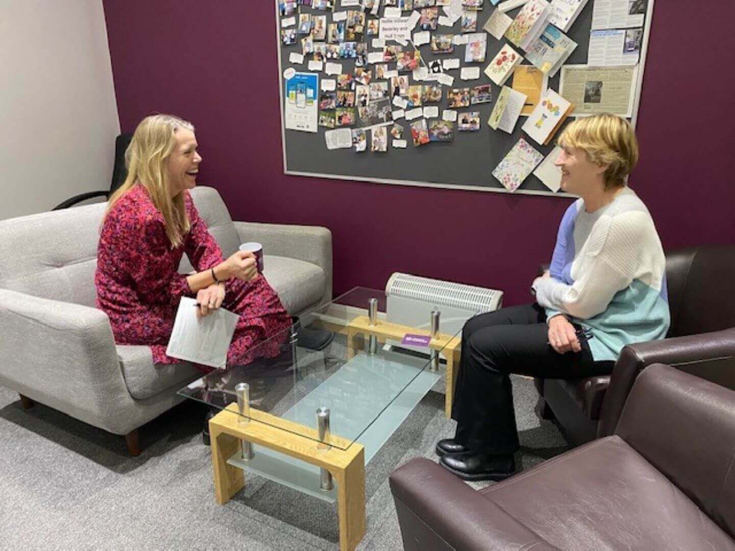 Two women sit and chat in an office lounge, with a bulletin board full of photos and notes on the wall behind them. - Home Instead