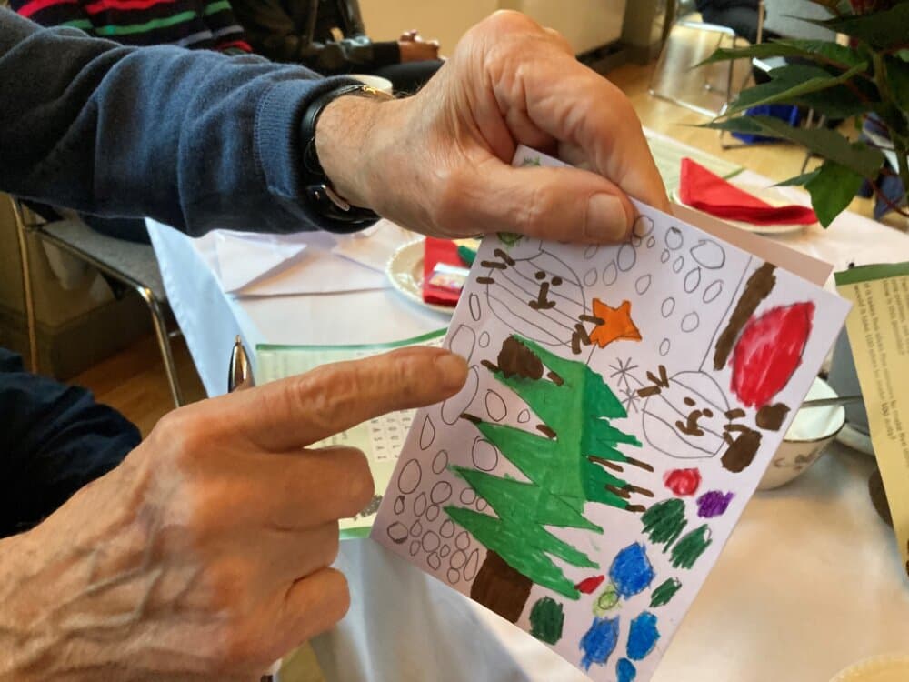 A person points at a colorful, hand-drawn holiday card featuring snowmen, a Christmas tree, and decorations. - Home Instead