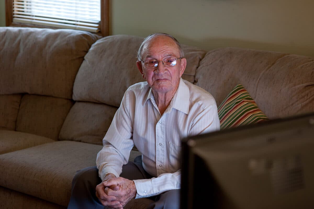 Elderly man with glasses, sitting on a couch, watching television in a living room. - Home Instead