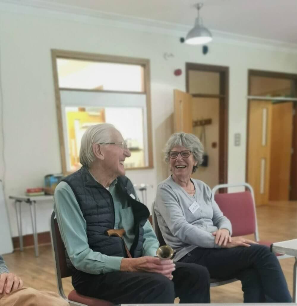Two older adults sit side by side in a bright room, smiling and laughing together. - Home Instead