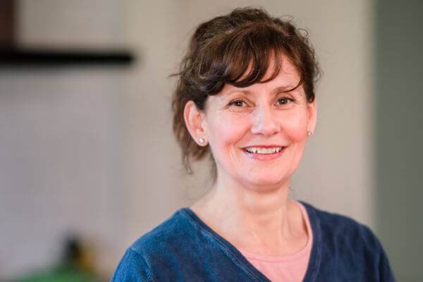 Smiling woman with wavy brown hair wearing a blue top, standing in a softly lit indoor setting. - Home Instead