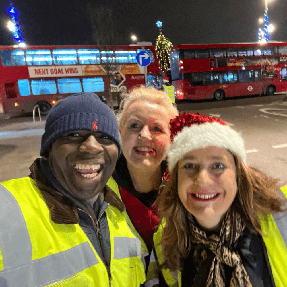 Three people in high-visibility jackets smile at night in front of double-decker buses; one wears a Santa hat. - Home Instead
