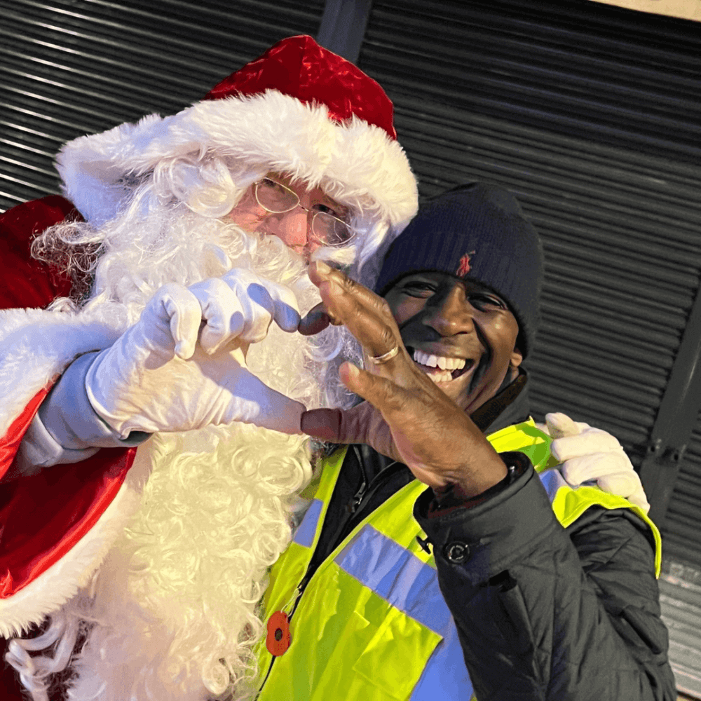 Santa Claus and a smiling person in a reflective jacket make heart shapes with their hands together. - Home Instead