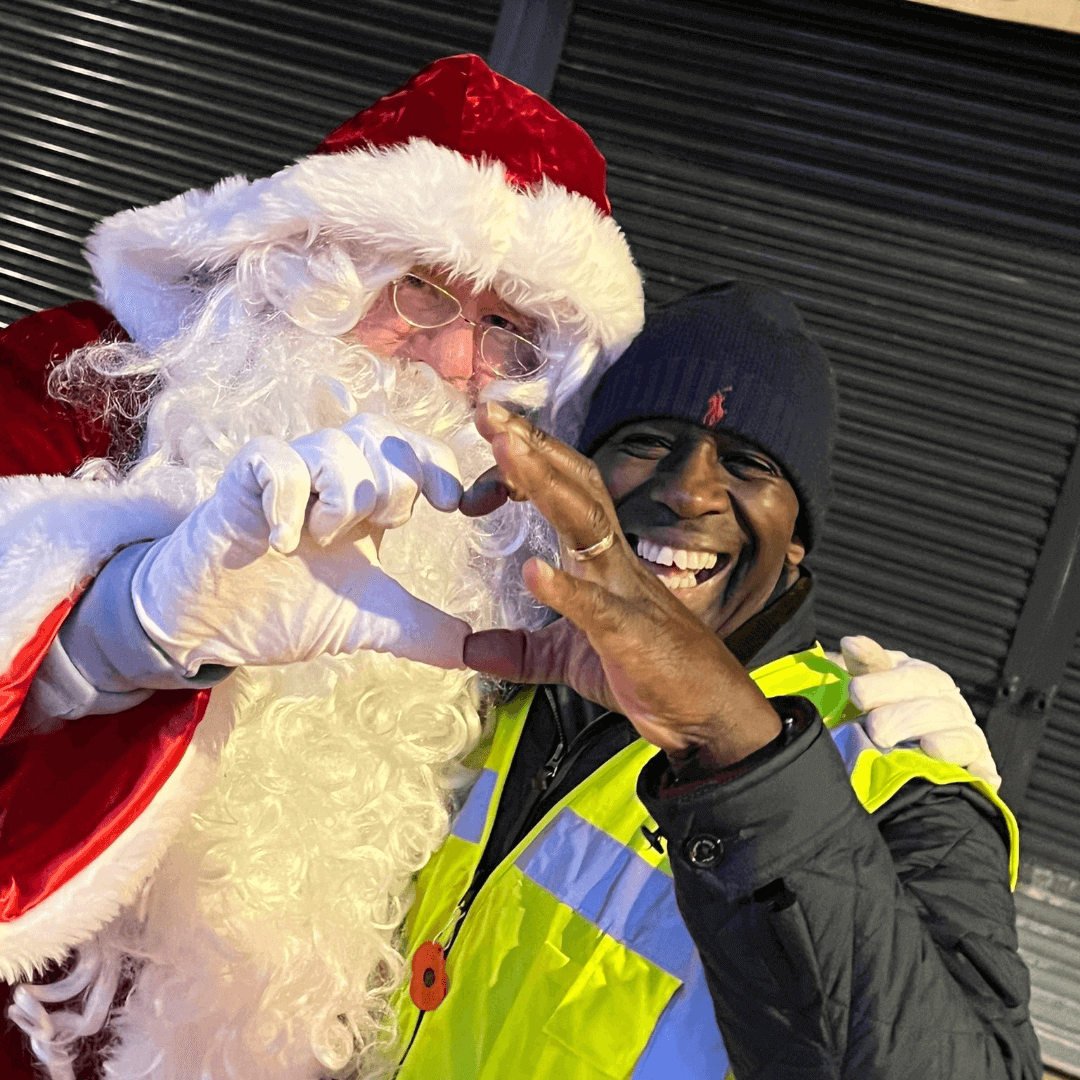 Santa Claus and a smiling person in a reflective jacket make heart shapes with their hands together. - Home Instead