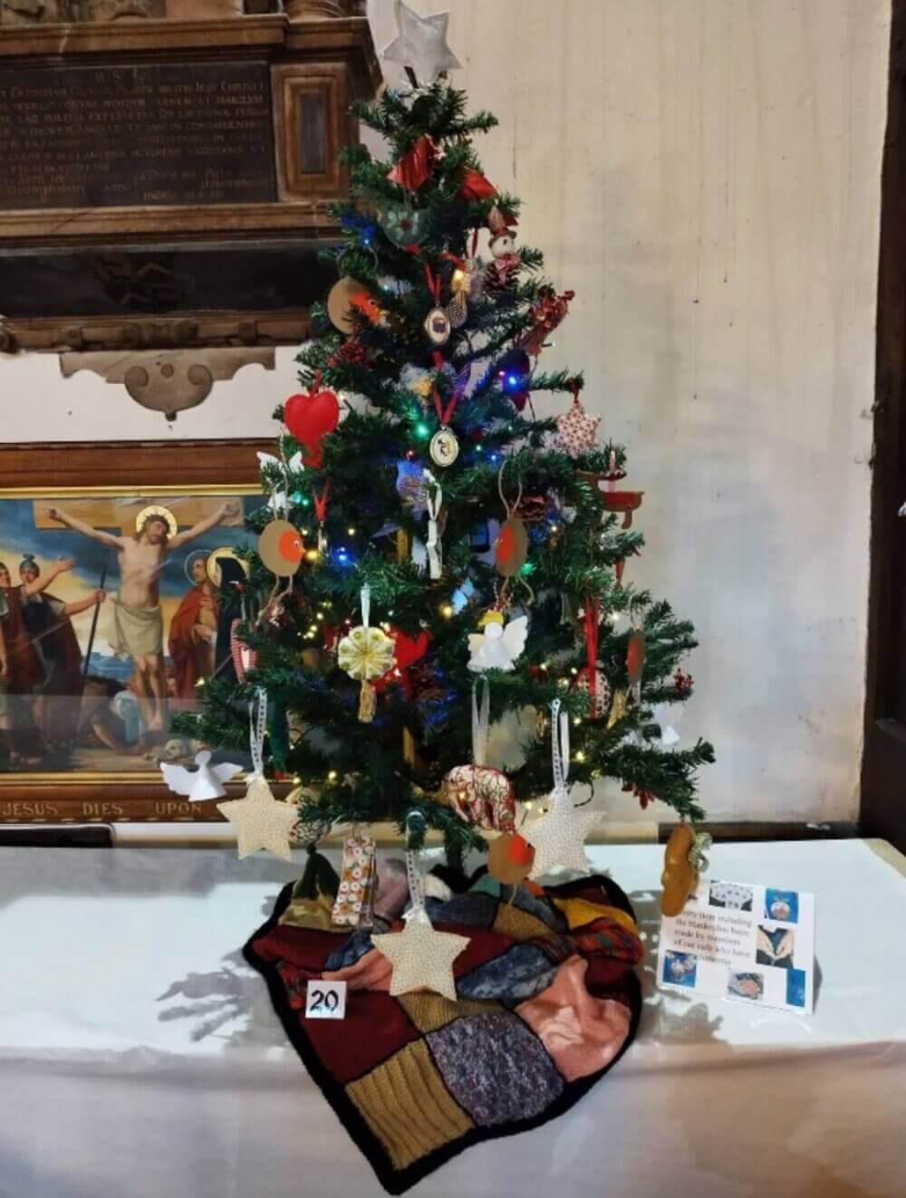 Decorated Christmas tree with lights and ornaments on a table, with a quilt and small items at its base. - Home Instead