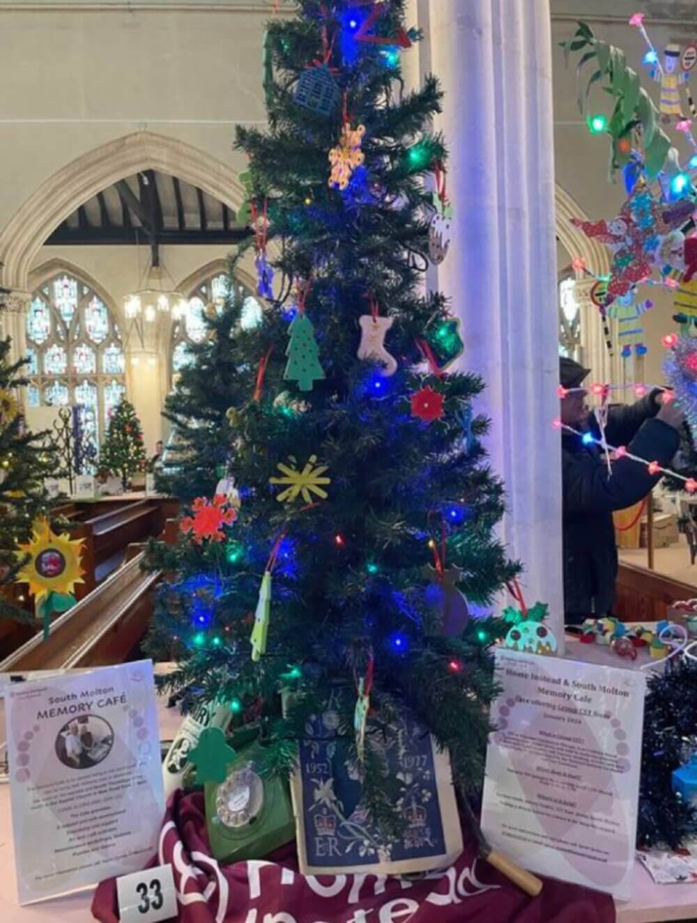 A decorated Christmas tree with lights and ornaments inside a church, surrounded by informational posters and flyers. - Home Instead