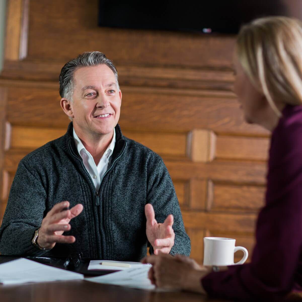 Two people seated at a table, engaged in a conversation, one gesturing with their hands. - Home Instead