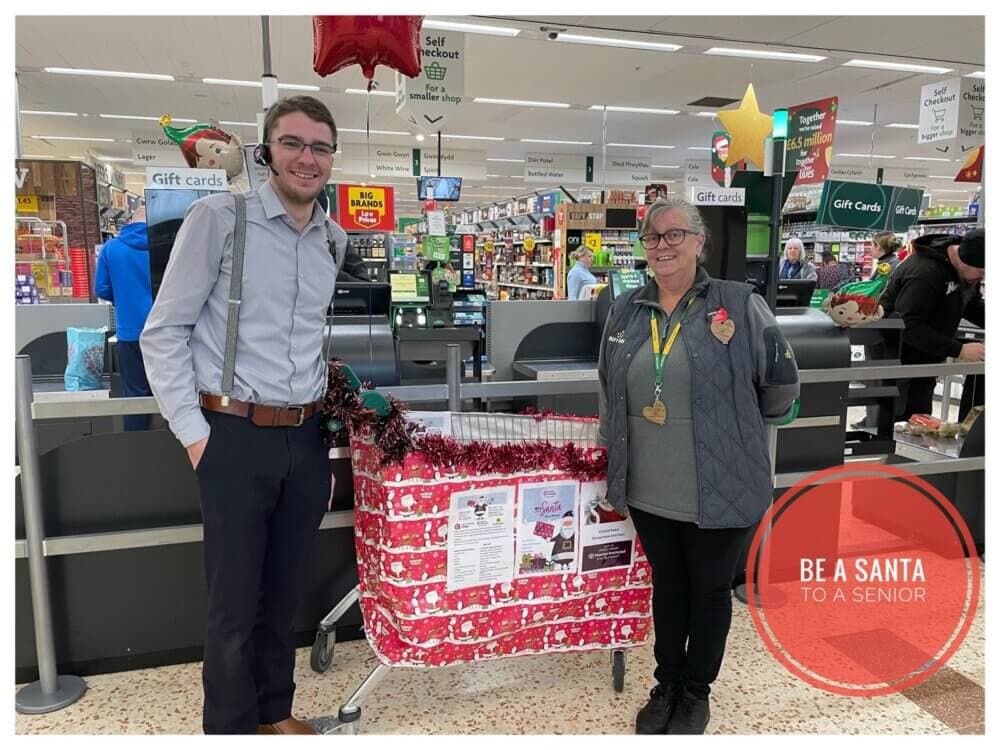 Two people standing beside a wrapped shopping cart with "Be a Santa to a Senior" sign in a store. - Home Instead