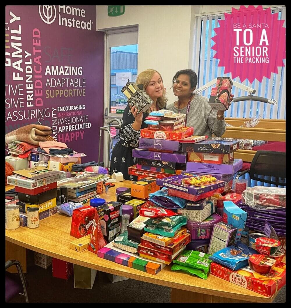 Two women smile while holding snacks next to a table full of stacked food items for a "Be a Santa to a Senior" event. - Home Instead