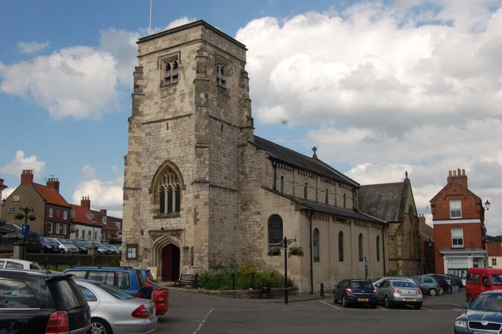 Stone church with a square tower stands at the center of a parking lot, surrounded by buildings and parked cars. - Home Instead