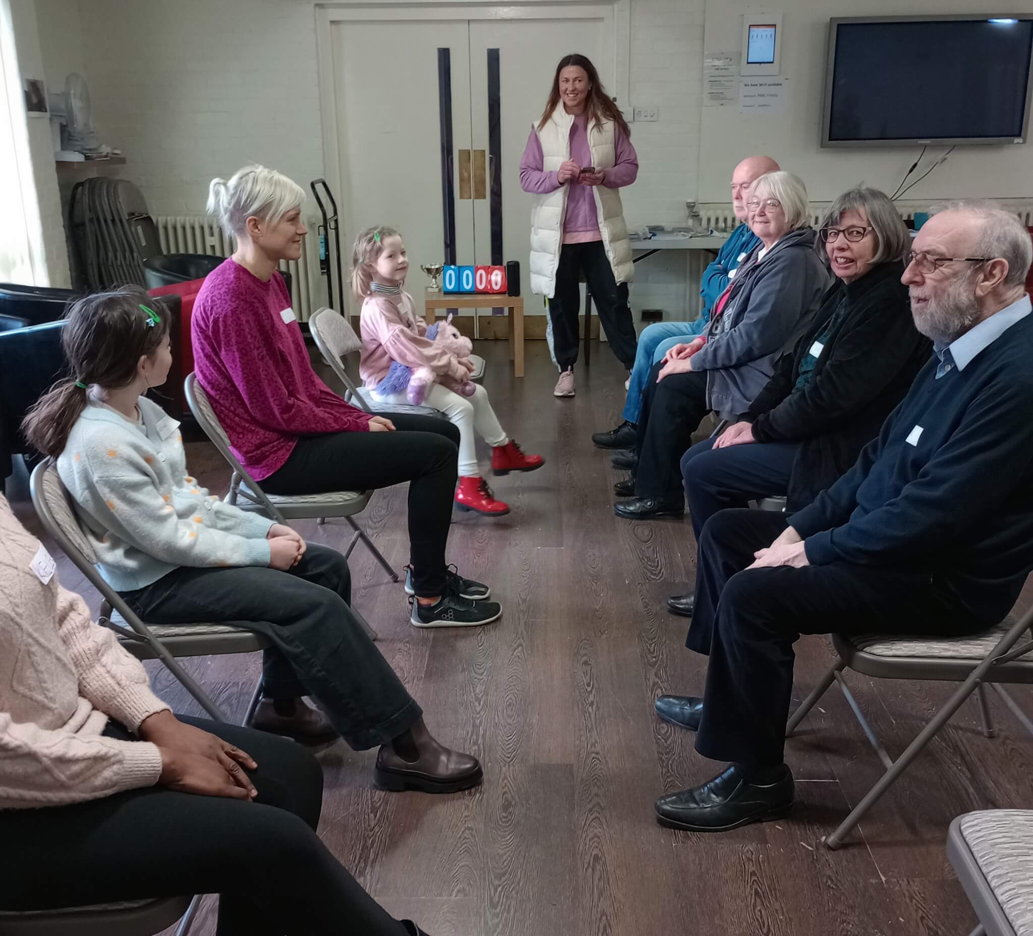 A group of people, including children and adults, sit in a semi-circle in a room, engaging in an activity. - Home Instead