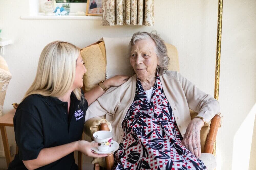 A caregiver smiles and holds a teacup for an elderly woman sitting in a chair in a cozy room. - Home Instead