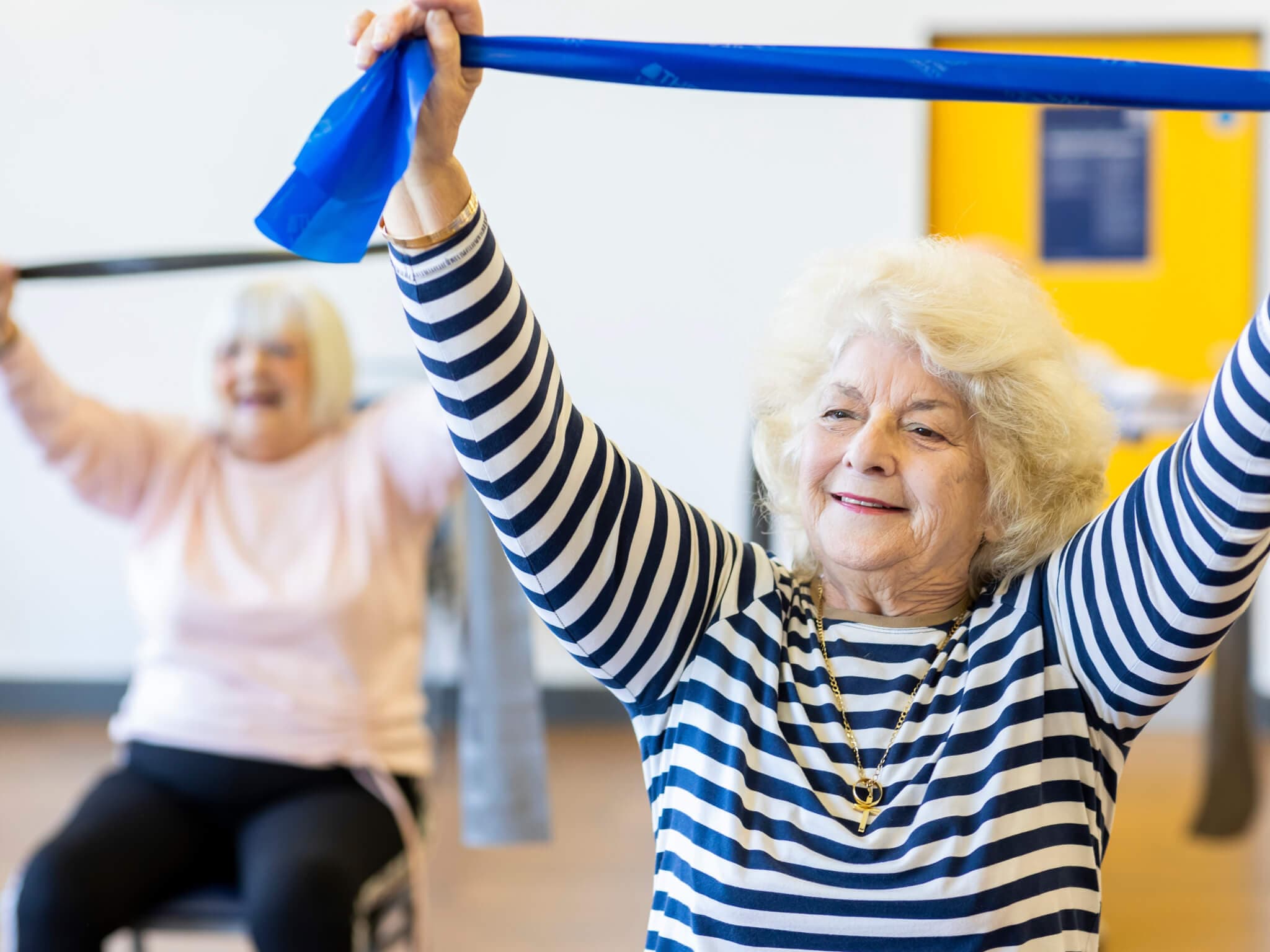 Two elderly women joyfully exercise with resistance bands in a bright room, with one in focus wearing a striped shirt. - Home Instead